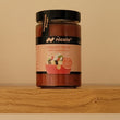 A glass jar of Nicola Tomato Frito with vegetables, featuring a black lid and a pinkish-brown label, sitting on a wooden shelf against a beige wall.