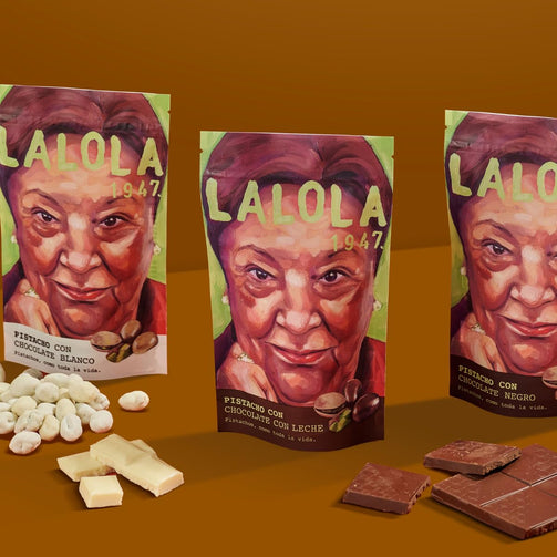 Three stand-up pouches of Lalola chocolate are arranged on a warm brown background, with loose white chocolate pieces on the left and dark chocolate pieces on the right. The pouches feature a painted portrait of a woman and the brand name Lalola 1947.