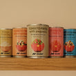 Four small cans of food products lined up on a wooden shelf against a plain beige wall. The cans have colorful labels, including yellow, peach, white, pink, and blue, with the brand name 'FF Foods' visible on each can.Nicola tomato frito with peppers