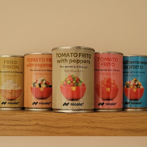 Four small cans of food products lined up on a wooden shelf against a plain beige wall. The cans have colorful labels, including yellow, peach, white, pink, and blue, with the brand name 'FF Foods' visible on each can.Nicola tomato frito with peppers 