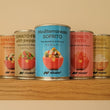 Six cans of Nicolo brand Mediterranean sofrito are lined up on a wooden shelf against a warm beige wall. The center can is light blue with a picture of a salad, while the surrounding cans are in orange, green, red, and yellow packaging, all featuring the Nicolo brand logo.