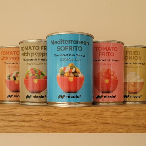Six cans of Nicolo brand Mediterranean sofrito are lined up on a wooden shelf against a warm beige wall. The center can is light blue with a picture of a salad, while the surrounding cans are in orange, green, red, and yellow packaging, all featuring the Nicolo brand logo.