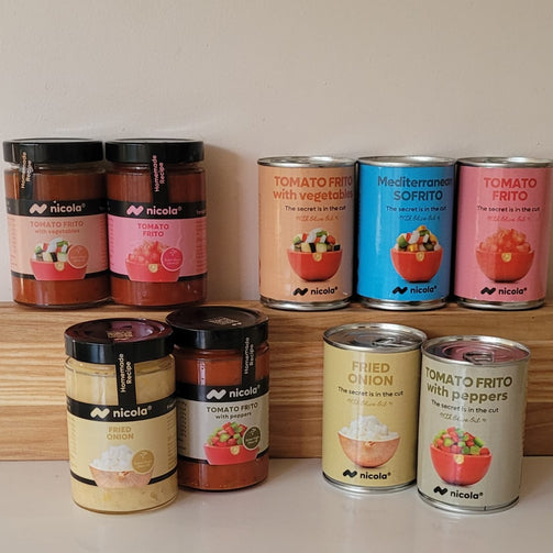 A collection of Nicole's Mediterranean and spanish salsa tomato frto, including jars and cans of tomato-based dishes, arranged on a wooden shelf and a white surface against a plain beige wall.